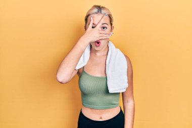 Spor kıyafetleri ve havlu giymiş genç sarışın kız şok içinde yüzünü ve gözlerini kapayarak utangaç ifadesiyle parmaklarının arasından bakıyor.. 