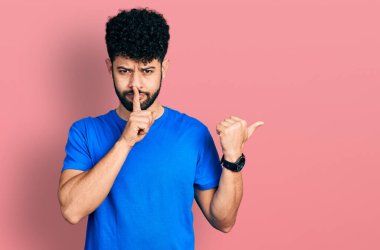 Sakallı, günlük mavi tişörtlü genç bir Arap sessiz olmak istiyor. Parmakları dudaklarında, elleri yanda. sessizlik ve gizli konsept. 