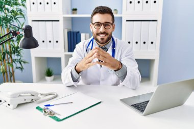 Young hispanic man wearing doctor uniform using laptop sitting at clinic