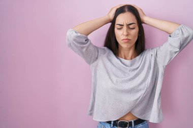 Genç esmer kadın pembe arka planda duruyor. Baş ağrısı, çaresiz ve stresli. Çünkü acı ve migren ağrısı var. Eller başının üstünde.. 