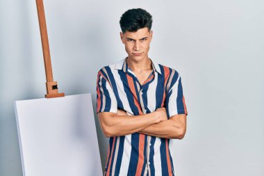 Young hispanic man standing close to empty canvas skeptic and nervous, disapproving expression on face with crossed arms. negative person. 