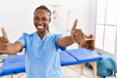 Black woman with braids working at pain recovery clinic approving doing positive gesture with hand, thumbs up smiling and happy for success. winner gesture. 