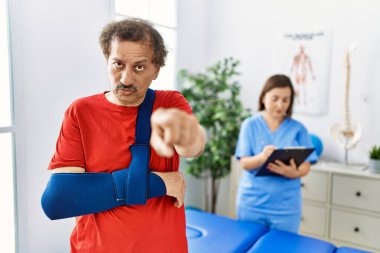 Southeast asian man wearing wearing arm on sling at rehabilitation clinic pointing with finger to the camera and to you, confident gesture looking serious 