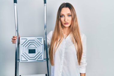 Genç sarışın kız merdiven tutuyor. Düşünme tavrı ve ayık yüz ifadesi. Kendine güveniyor gibi görünüyor. 