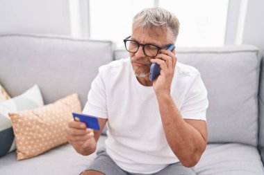 Orta yaşlı, kır saçlı, akıllı telefon ve kredi kartıyla konuşan adam evdeki kanepede oturuyor.