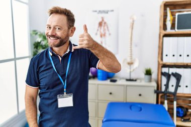 Middle age physiotherapist man working at pain recovery clinic smiling doing phone gesture with hand and fingers like talking on the telephone. communicating concepts. 
