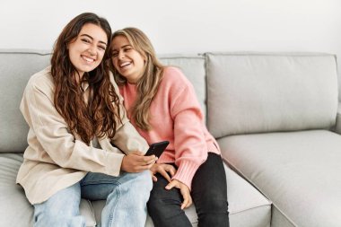 Genç çift evde koltukta oturmuş akıllı telefon kullanarak mutlu bir şekilde gülümsüyor..