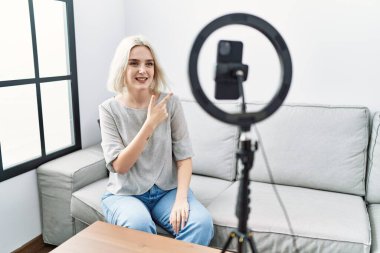 Young caucasian woman recording vlog tutorial with smartphone at home cheerful with a smile of face pointing with hand and finger up to the side with happy and natural expression on face 