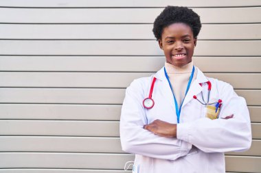 Doktor üniforması giyen Afrikalı Amerikalı kadın caddede kollarını kavuşturmuş duruyor.