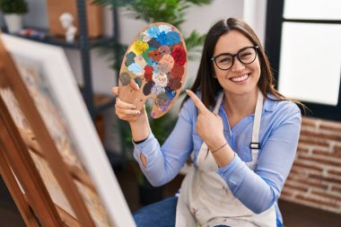 Sanat stüdyosunda boyacı paleti tutan genç esmer kadın mutlu bir şekilde elini ve parmağını gösterdi. 