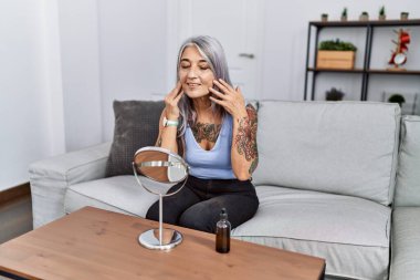 Orta yaşlı, kır saçlı, cilt bakımı gören bir kadın. Evdeki kanepede oturuyor.