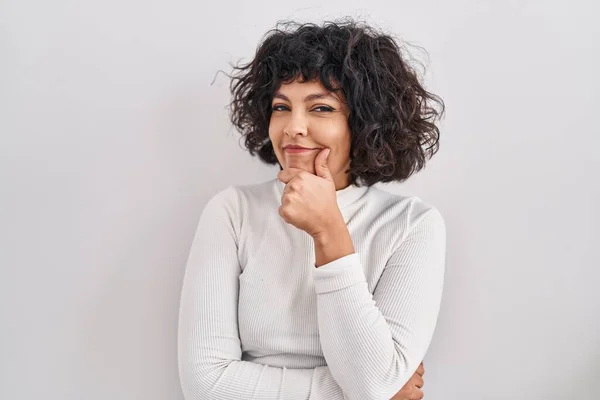 Hispanic woman with curly hair standing over isolated background looking confident at the camera smiling with crossed arms and hand raised on chin. thinking positive.