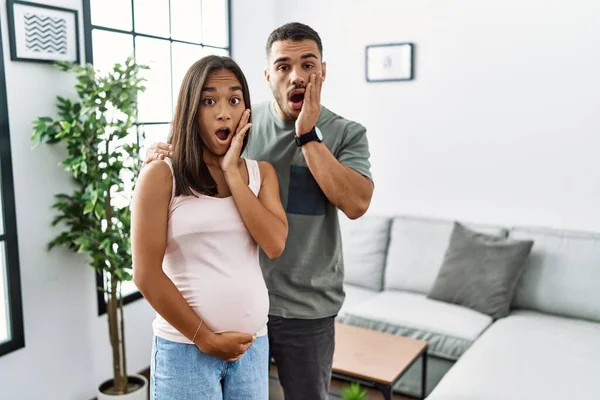 Young interracial couple expecting a baby, touching pregnant belly afraid and shocked, surprise and amazed expression with hands on face