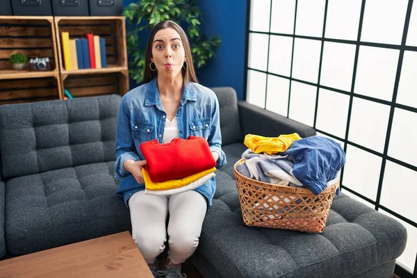 Hispanic woman holding folded laundry after ironing making fish face with mouth and squinting eyes, crazy and comical.