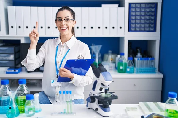 Young brunette woman working at scientist laboratory smiling amazed and surprised and pointing up with fingers and raised arms.
