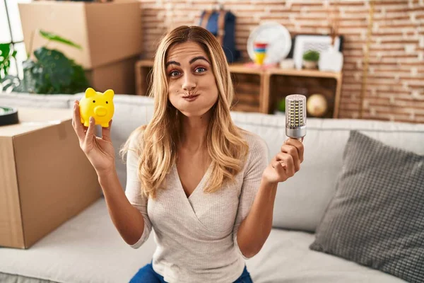 Young blonde woman holding led bulb and piggy bank puffing cheeks with funny face. mouth inflated with air, catching air.