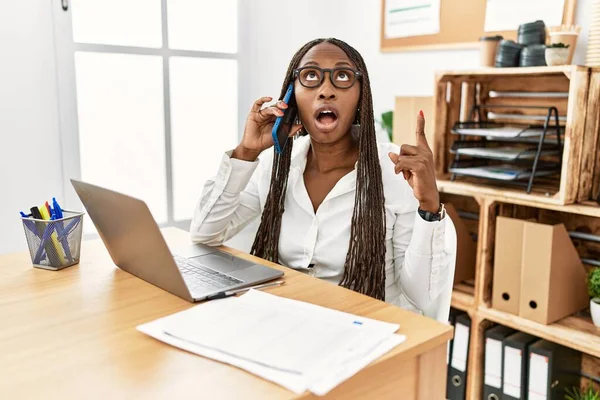 Black woman with braids working at the office speaking on the phone amazed and surprised looking up and pointing with fingers and raised arms.