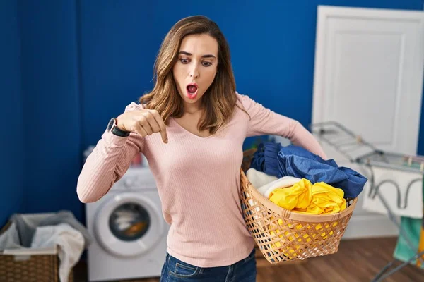 Young woman holding laundry basket pointing down with fingers showing advertisement, surprised face and open mouth