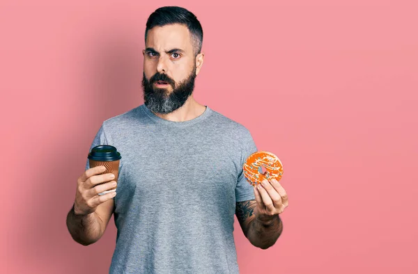 Hispanic man with beard eating doughnut and drinking coffee in shock face, looking skeptical and sarcastic, surprised with open mouth