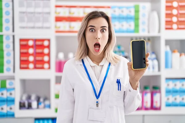 Young beautiful woman working at pharmacy drugstore showing smartphone screen scared and amazed with open mouth for surprise, disbelief face