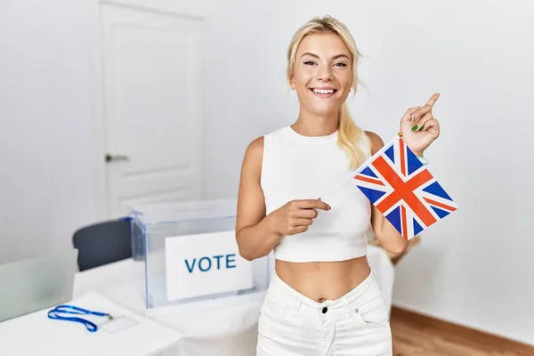 Young caucasian woman at political campaign election holding uk flag smiling happy pointing with hand and finger to the side