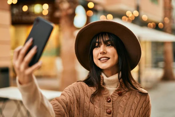 Kışlık şapkalı esmer bir kadın şehirde akıllı telefonuyla selfie çekiyor.