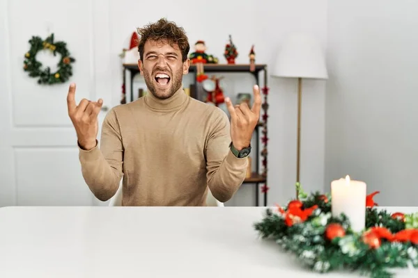 クリスマスの装飾によってテーブルの上に座ってひげを生やした若いハンサムな男が手を上げて岩のシンボルをやって狂気の表情で叫んでいます 音楽スター 重い音楽の概念は — ストック写真