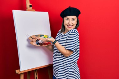 Middle age hispanic woman standing drawing with palette by painter easel stand pointing aside with hands open palms showing copy space, presenting advertisement smiling excited happy 
