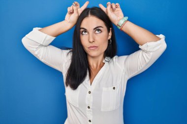 Mavi arka planda dikilen genç İspanyol kadın parmaklarıyla boynuz gibi komik hareketler yapıyor. 