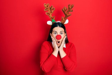 Young hispanic woman wearing deer christmas hat and red nose afraid and shocked, surprise and amazed expression with hands on face 