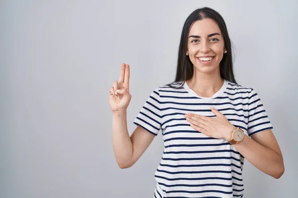 Young brunette woman wearing striped t shirt smiling swearing with hand on chest and fingers up, making a loyalty promise oath