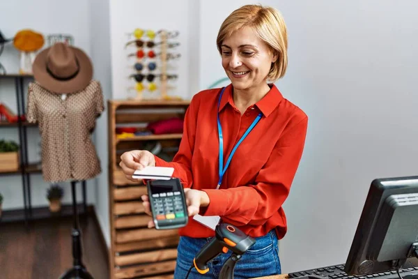 Middle age blonde woman smiling confident using credit card and data phone at clothing store