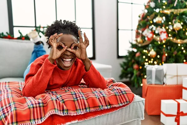 Young african american woman lying on the sofa by christmas tree doing ok gesture like binoculars sticking tongue out, eyes looking through fingers. crazy expression.