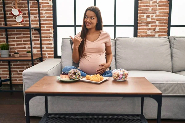 Mujer Embarazada Joven Comiendo Dulces Casa Apuntando Pulgar Hacia Lado —  Fotos de Stock