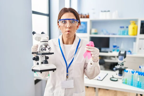 Young brunette woman working at scientist laboratory holding pink ribbon making fish face with mouth and squinting eyes, crazy and comical.