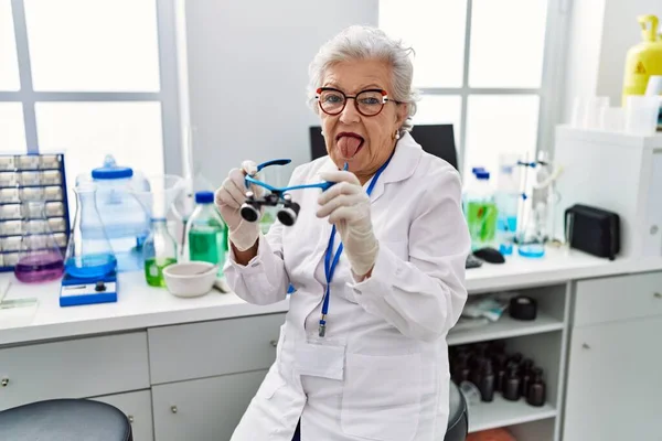 科学研究所で働く白髪のシニア女性が舌を突き出す虫眼鏡を使って面白い表情で幸せ — ストック写真