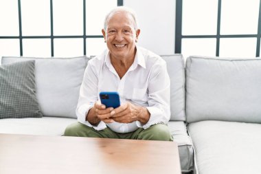 Kıdemli adam evde akıllı telefon kullanarak kendinden emin gülümsüyor.