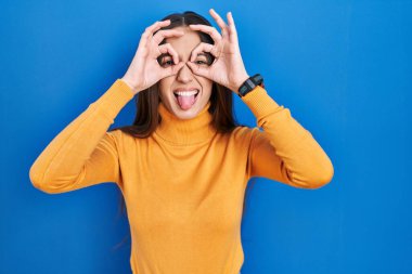 Mavi arka planda duran genç esmer kadın dürbün gibi jest yapıyor. Dürbün dilini çıkarıyor, gözler parmaklarından bakıyor. çılgın ifade. 