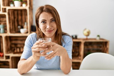 Orta yaşlı beyaz bir kadın evdeki masada oturmuş su içiyor..