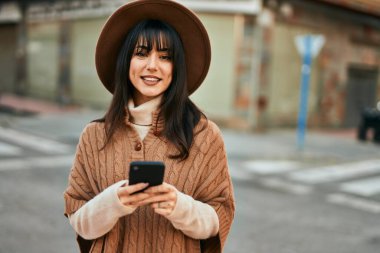 Kışlık şapkalı esmer kadın, şehirde akıllı telefon kullanarak gülümsüyor.