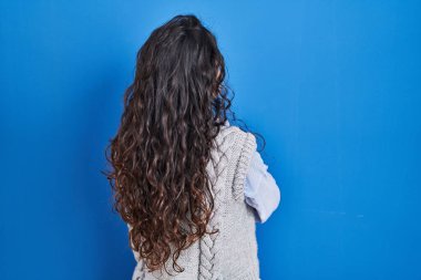 Genç esmer kadın mavi arka planda dikiliyor geriye bakıyor ve kollarını kavuşturuyor. 