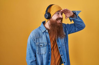Caucasian man with long beard listening to music using headphones very happy and smiling looking far away with hand over head. searching concept. 