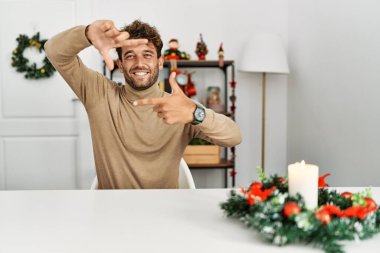 Masada sakallı genç yakışıklı bir adam Noel süslemesi ile oturuyor gülümsüyor ve elleri ve parmakları gülen yüzüyle çerçeve yapıyor. yaratıcılık ve fotoğrafçılık kavramı. 