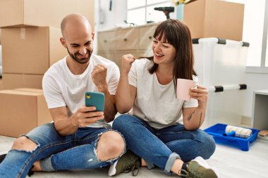 Genç İspanyol çift yeni evinde akıllı telefon kullanarak mutlu bir şekilde gülümsüyor..
