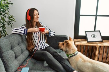 Genç İspanyol kadın müzik dinliyor ve evdeki köpekle kanepede oturup kahve içiyor.