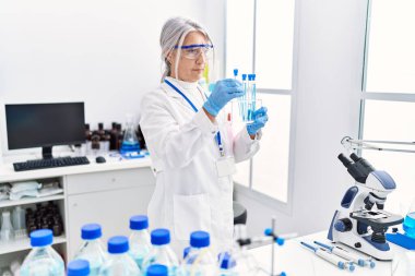 Orta yaşlı, gri saçlı, bilim adamı üniforması giyen bir kadın laboratuarda dizüstü bilgisayar kullanarak test tüpü tutuyor.