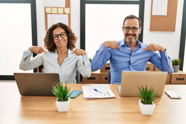 Middle age hispanic woman and man sitting with laptop at the office looking confident with smile on face, pointing oneself with fingers proud and happy. 