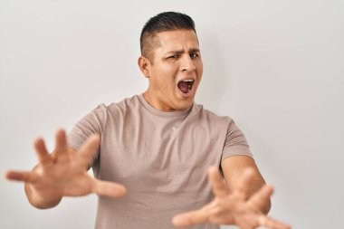Hispanic young man standing over white background afraid and terrified with fear expression stop gesture with hands, shouting in shock. panic concept. 