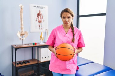 Fizyoterapi kliniğinde çalışan genç İspanyol bir kadın basketbol topunu depresif ve endişeli tutuyor. Kızgın ve korkmuş bir şekilde ağlıyor. hüzünlü ifade. 