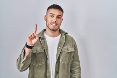 İzole edilmiş arka planda duran genç İspanyol adam bir numarayı gösterip işaret ederken kendinden emin ve mutlu gülümsüyor.. 